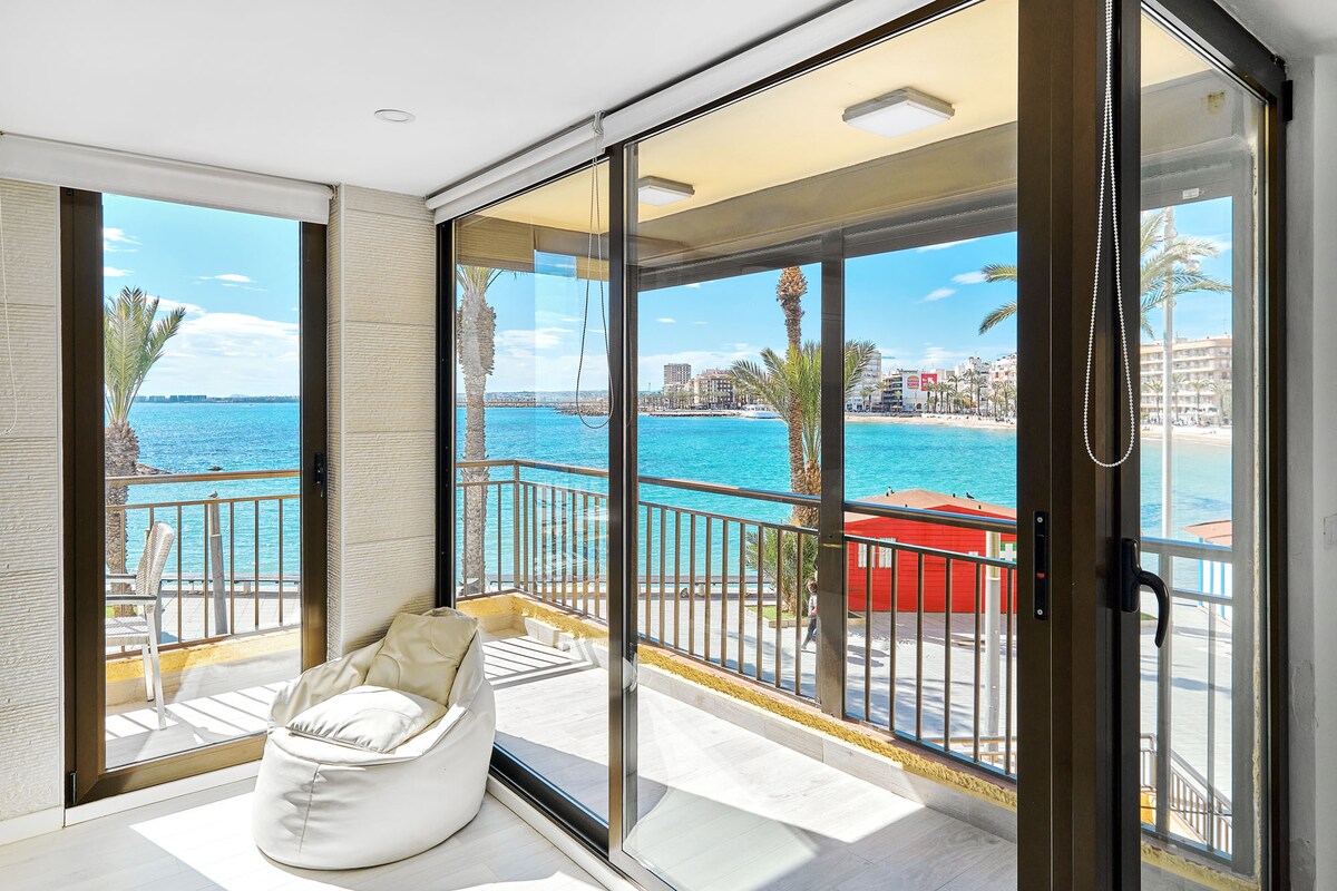 Large glass doors create a seamless transition between indoor and outdoor living, showcasing expansive views of Playa del Cura beach. A cozy bean bag chair sits on the light-colored flooring, inviting relaxation while palm trees and calm waters form a serene backdrop.