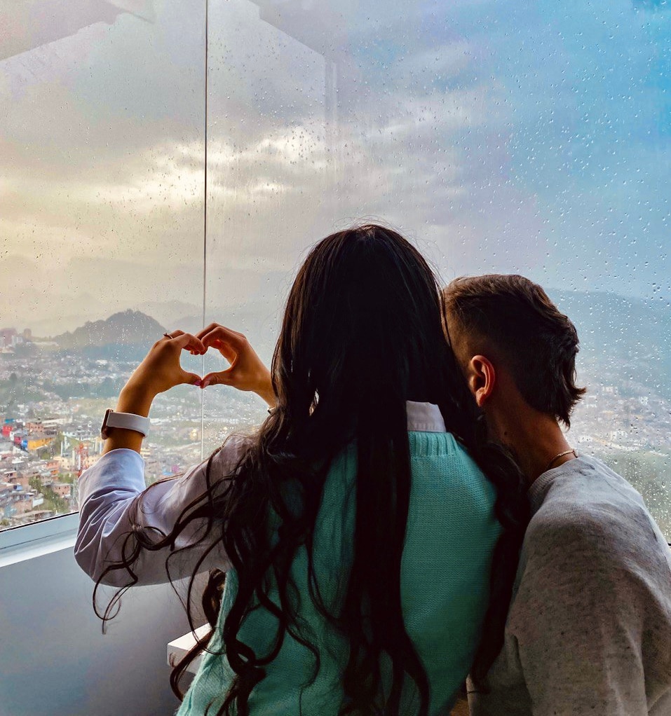 A couple is seen from behind, forming a heart shape with their hands. They are gazing out of a large window, showcasing a view of the city under a rainy sky. Raindrops are visible on the glass, adding a soft focus to the landscape beyond.