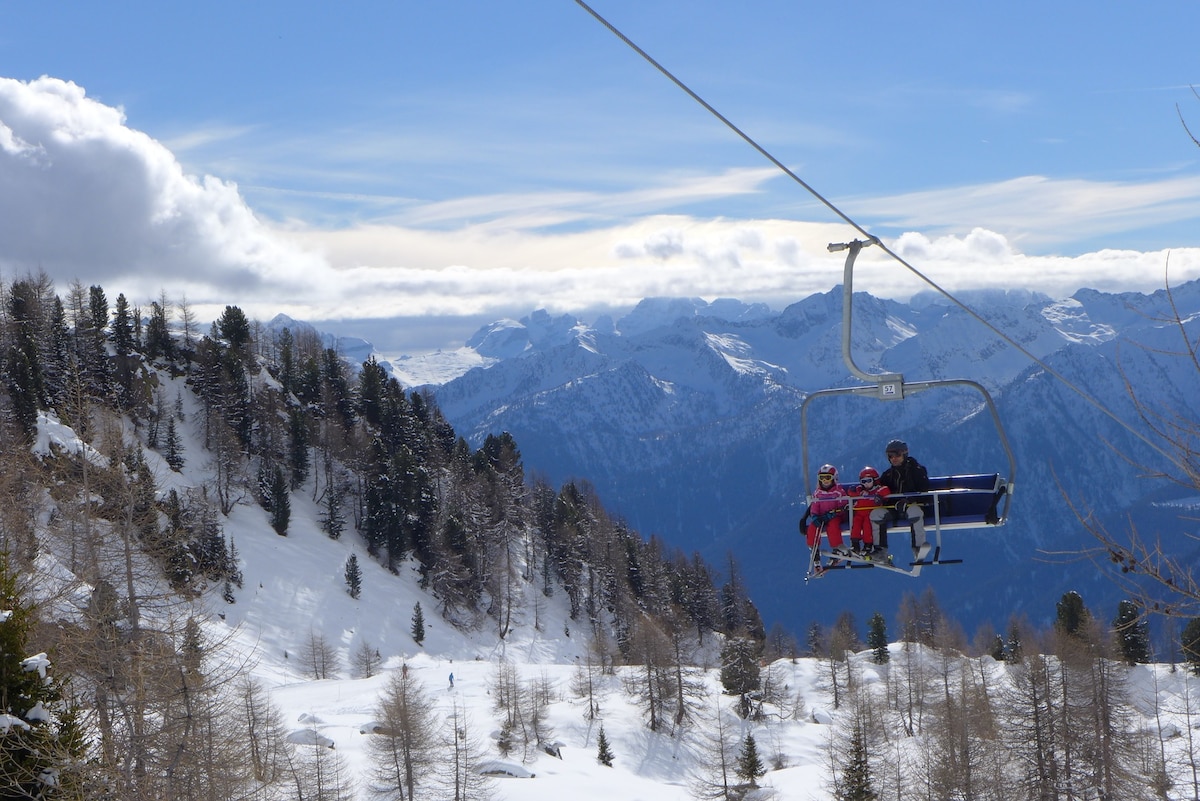 A ski lift carries three passengers uphill, surrounded by snow-covered mountains and evergreen trees. The expansive landscape reveals a variety of peaks under a partly cloudy sky, showcasing the serene beauty of the Dolomites.