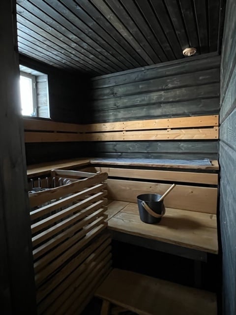 A wooden sauna interior is presented, featuring dark walls and a cedar ceiling. Benches made of light-colored wood line the space, complemented by a traditional sauna stove and an adjacent wooden bucket. A small window allows natural light to filter in.