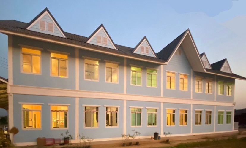A charming two-story building features a light blue exterior with multiple gabled rooftops. Warm light spills from the windows, illuminating the structure. Each floor showcases a series of evenly spaced windows, enhancing the overall design while providing a welcoming appearance.