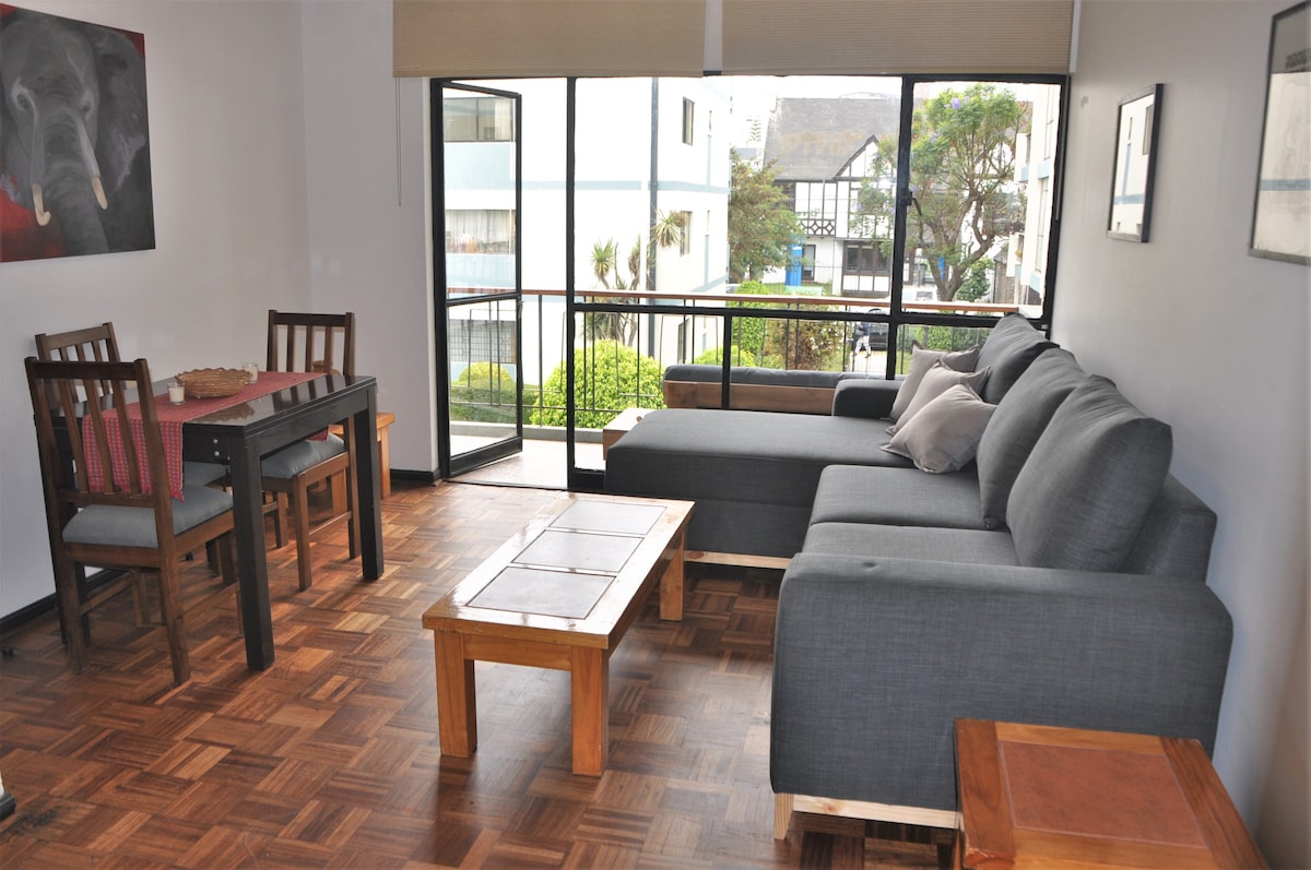 The living and dining area features a sectional sofa and a dining table set for four. Large windows allow natural light to fill the space, while providing views of the surrounding gardens and courtyard. The floor is adorned with wooden parquet, enhancing the room's warmth.