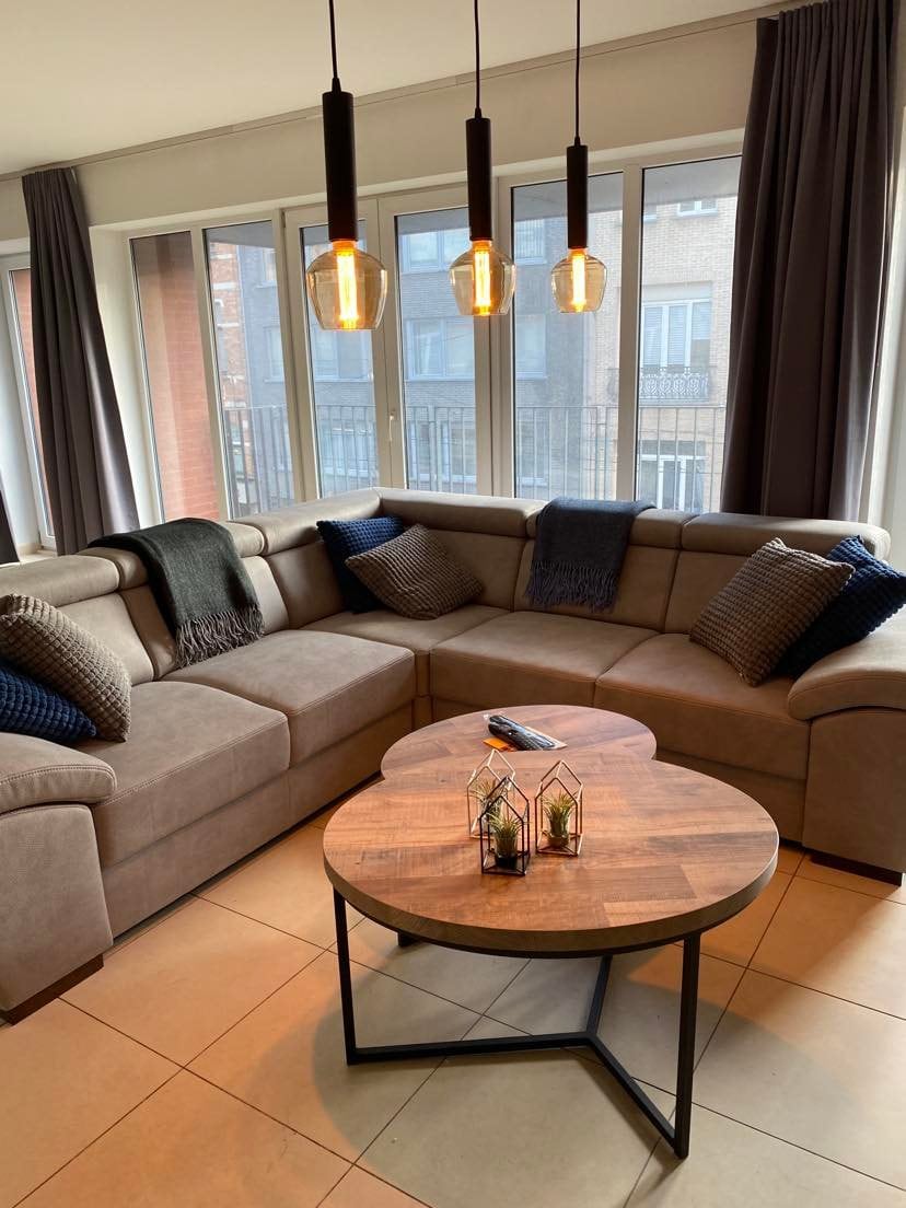 A sectional sofa is arranged in the corner, adorned with dark blue and textured throw pillows. A round wooden coffee table is placed in front, featuring decorative glass lanterns. Natural light streams in through large windows, dressed with floor-length gray curtains.