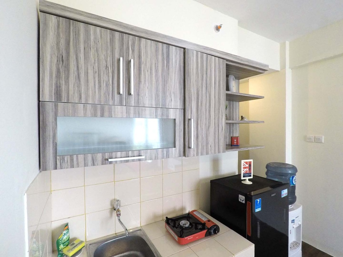A compact kitchen area features modern wooden cabinetry with a glass panel, providing storage for kitchen essentials. A circular sink sits alongside a gas stove, while a refrigerator is positioned nearby, creating a functional cooking space within the studio apartment.