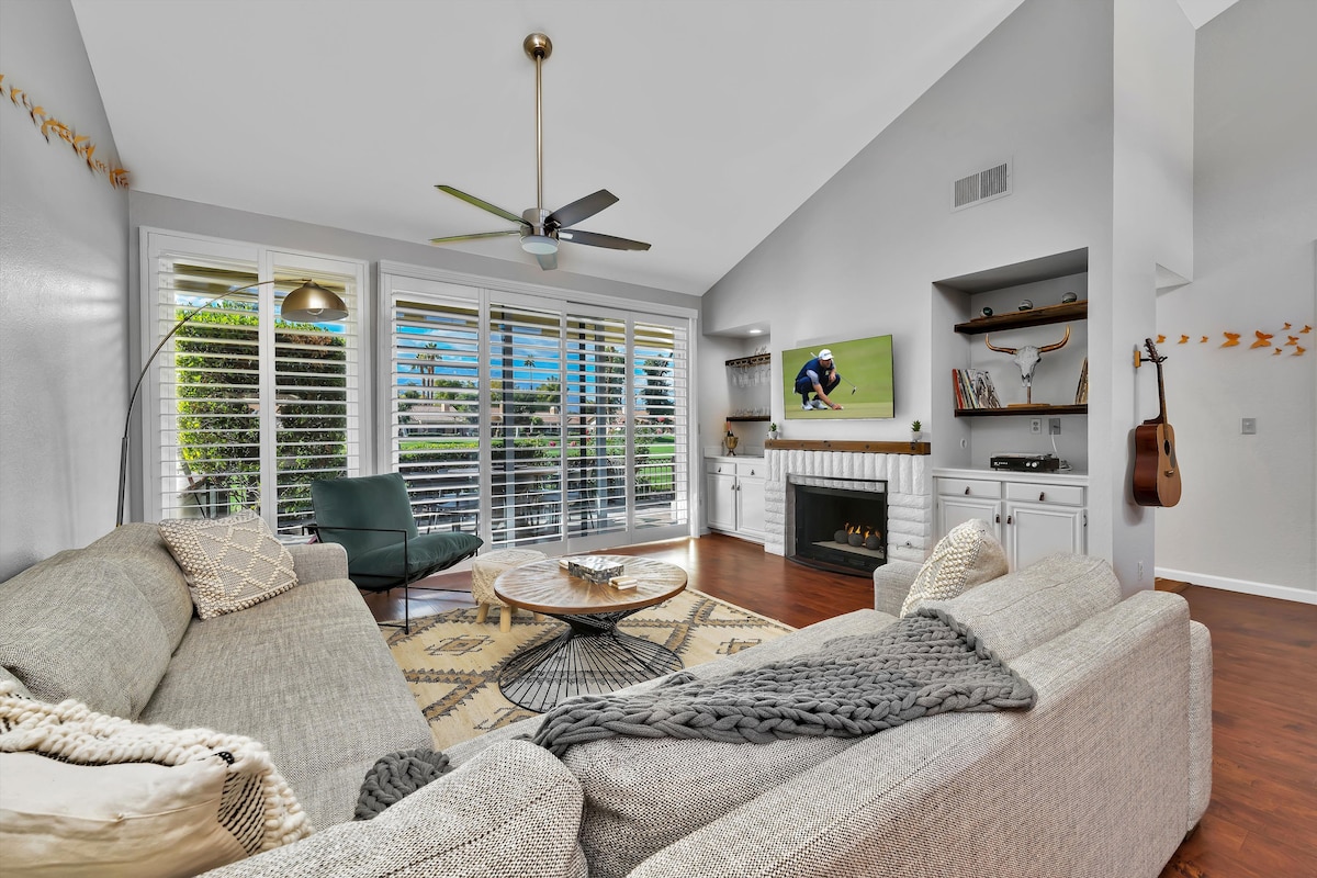 A spacious living room features a large L-shaped couch and a comfortable chair, complemented by a coffee table. Natural light flows through expansive windows adorned with shutters. A fireplace and a wall-mounted TV enhance the cozy ambience, while decorative accents complete the inviting space.