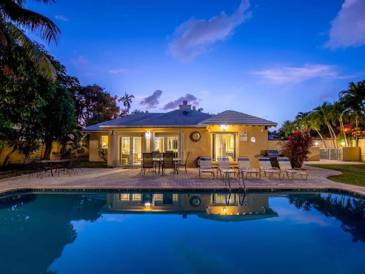 The exterior of a private mansion is reflected in a tranquil pool, framed by tropical landscaping. A spacious patio area features lounge chairs, with glass doors leading into the open living space. Warm evening light enhances the serene atmosphere.