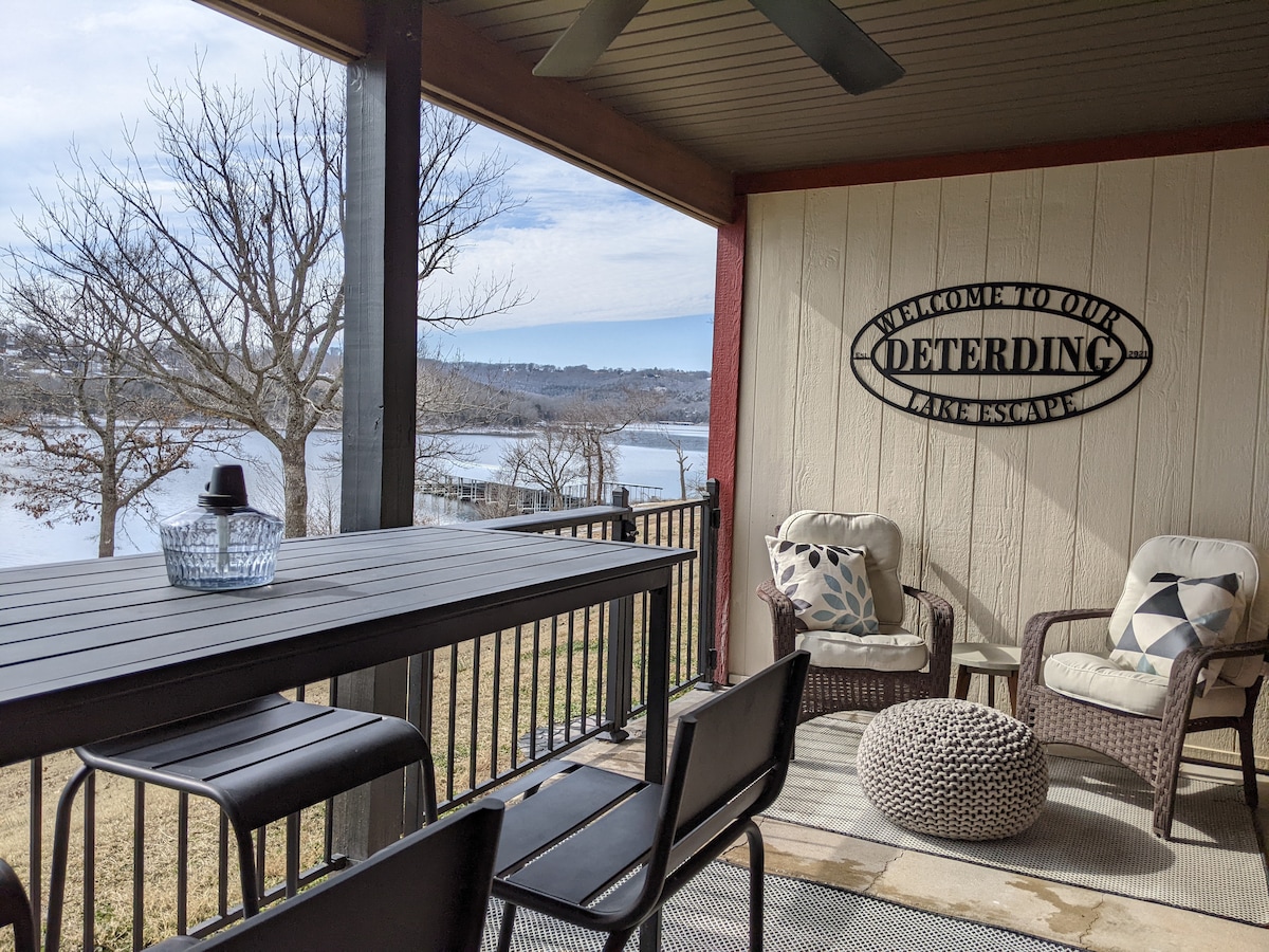 A covered patio offers a view of the lake, featuring a high-top table with stools and two comfortable chairs. A decorative welcome sign is mounted on the wall, and a woven rug is placed on the floor, adding a cozy touch to the outdoor space.