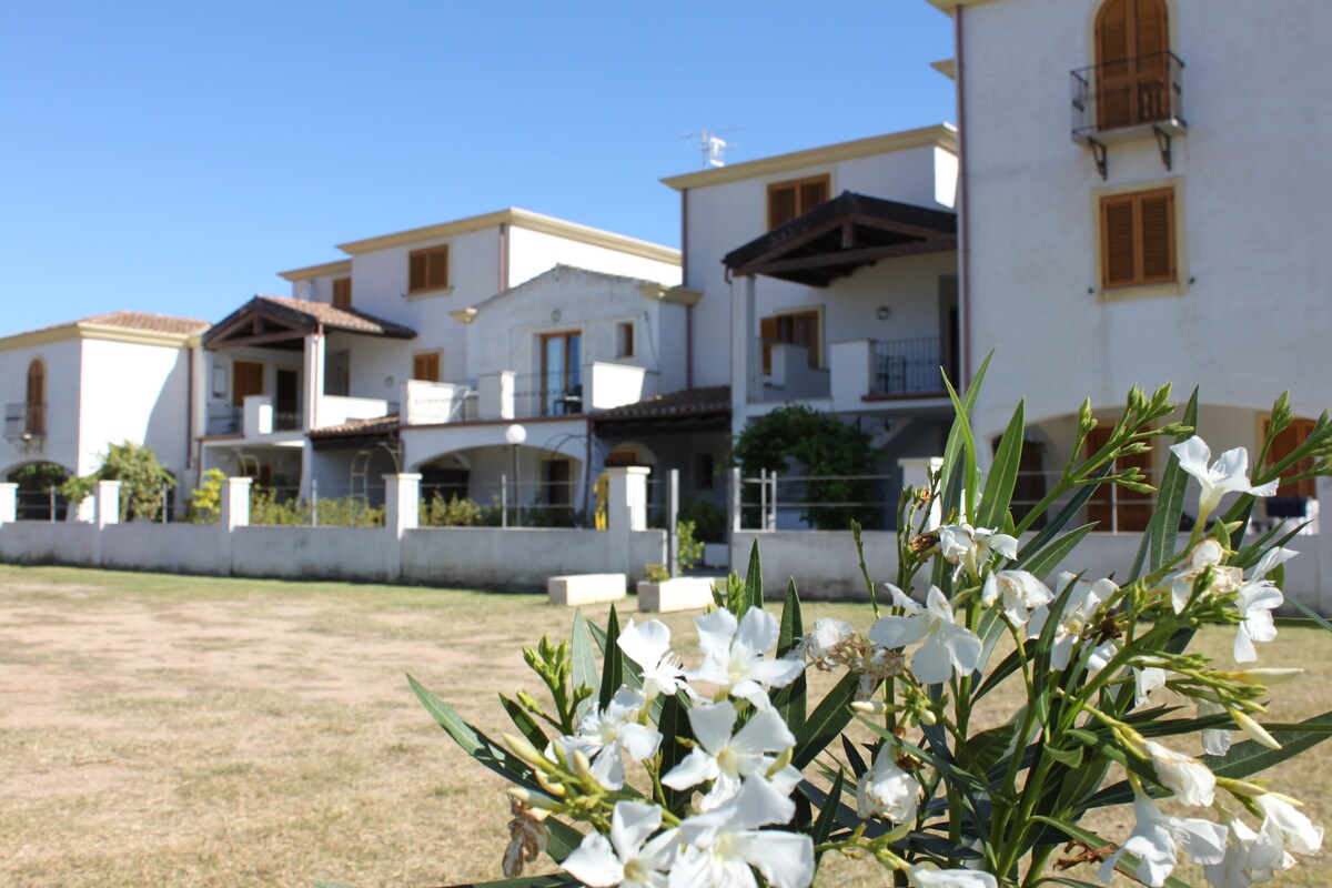 A modern collection of independent vacation homes is set against a clear blue sky. A garden with white flowering plants is visible in the foreground, while the three-story buildings feature balconies and varying architectural details, framed by well-kept grass in a serene environment.