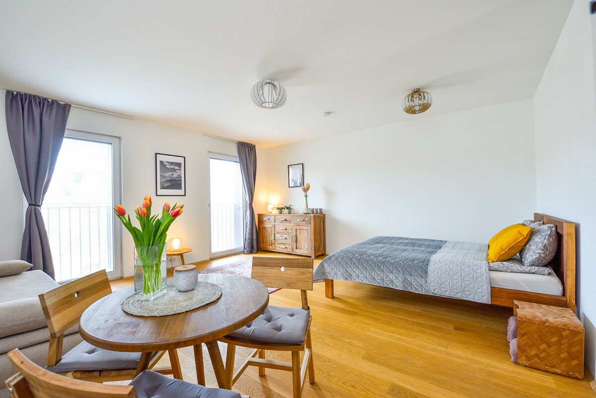A bright living space features large windows allowing natural light to fill the room. A spacious bed with decorative pillows is positioned alongside a stylish wooden dresser. A round dining table with four chairs is placed near the center, accompanied by a bouquet of tulips for a touch of color.