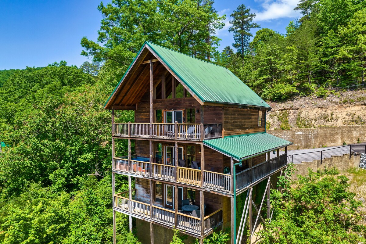 A three-story cabin is nestled among lush greenery, featuring a distinctive metal roof and wooden exterior. Expansive balconies wrap around the structure, providing views of the surrounding trees. The building is positioned on a slight slope, enhancing its secluded and elevated feel.
