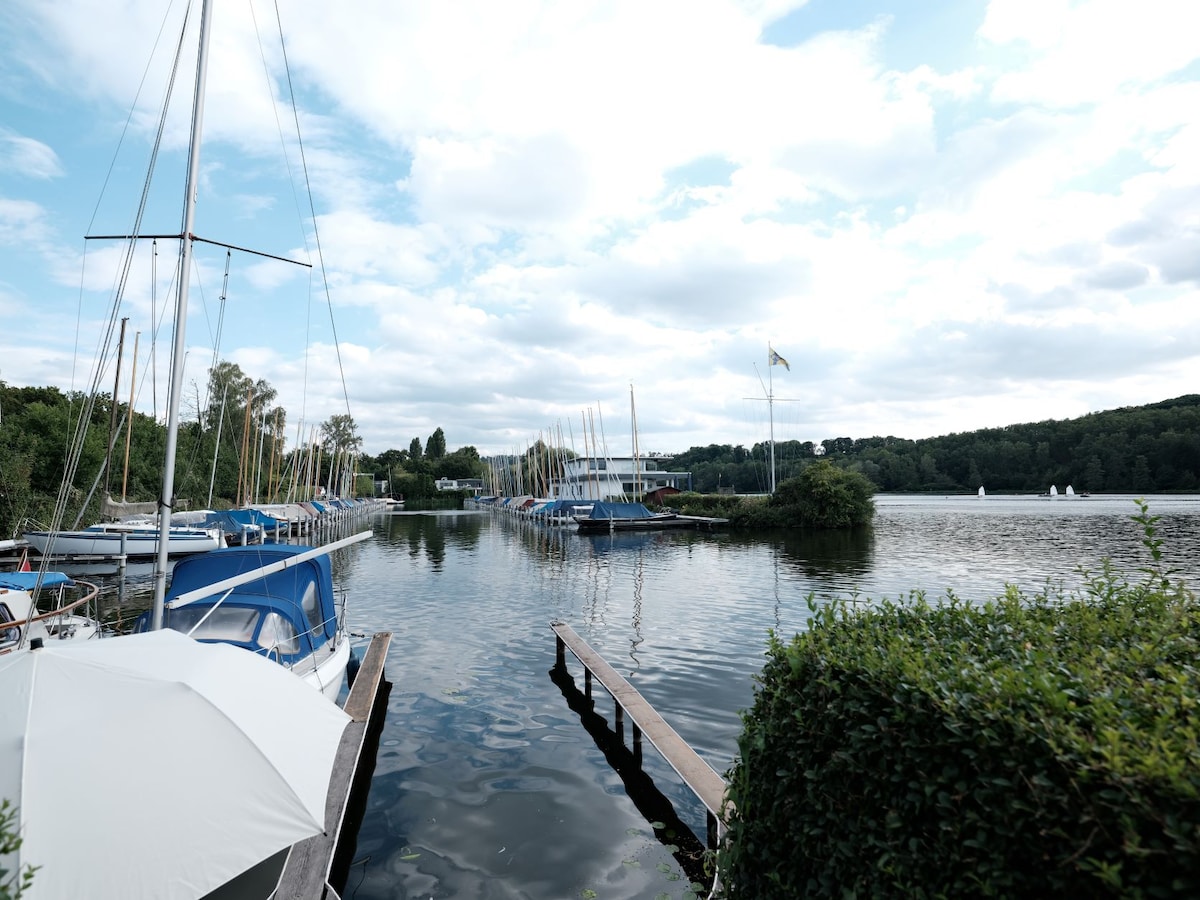 A serene waterfront scene features several boats moored along a wooden pier. The calm water reflects the sky, which is partly cloudy. Lush greenery borders the area, creating a tranquil atmosphere, while distant sailboats can be seen gliding gracefully across the water.
