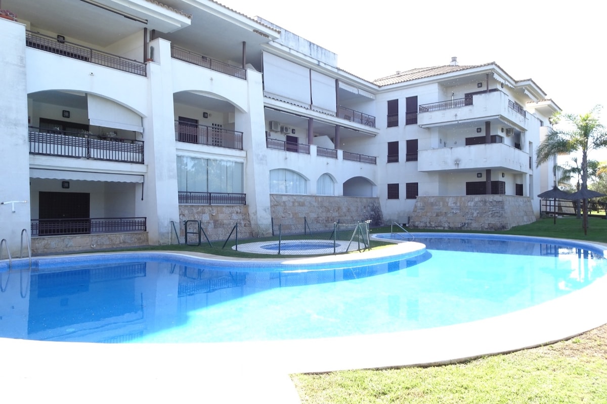 A refreshing swimming pool is centered within the landscaped grounds of the apartment complex, surrounded by grass and various architectural features. Large balconies are visible on the upper floors, with terraces providing shaded areas. The overall atmosphere suggests a well-maintained recreational space.
