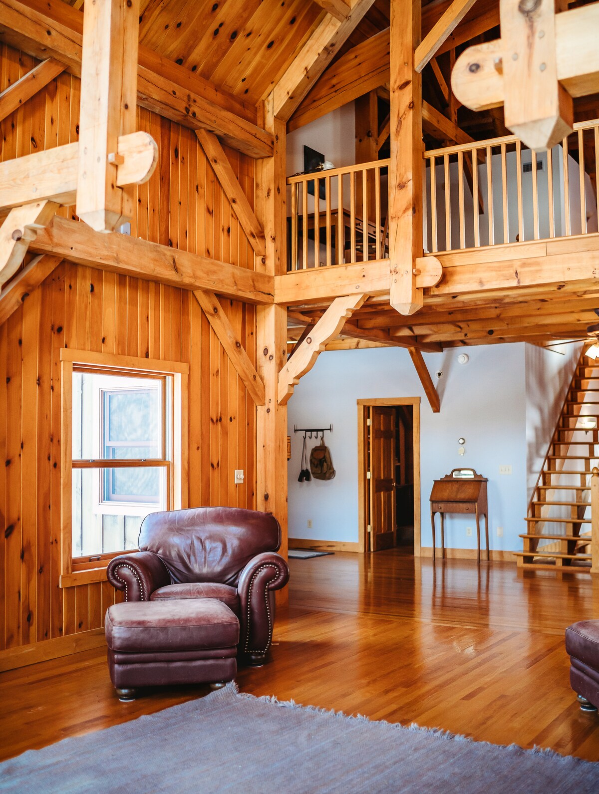 The interior features a spacious living area with high, exposed wooden beams and warm, polished hardwood floors. A leather armchair is positioned near a window, providing a cozy corner. A staircase leads to an upper level, enhancing the open and inviting design of the space.