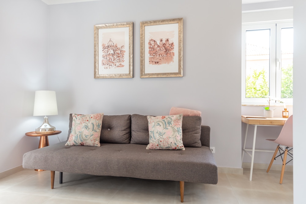 A neutral-toned living area features a low, modern sofa adorned with floral accent pillows. Two framed artworks hang on the wall above, complementing the light color scheme. A small wooden table with a lamp is positioned nearby, alongside a desk with a chair set by the window.
