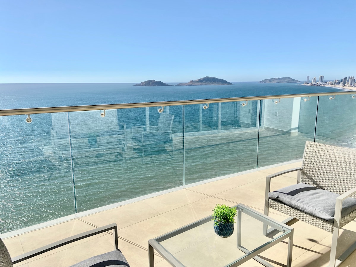 A spacious terrace is shown with a glass railing overlooking serene ocean waters. Two contemporary chairs and a glass-top table are positioned nearby, complemented by a small decorative plant. In the background, the coastline and islands can be seen.