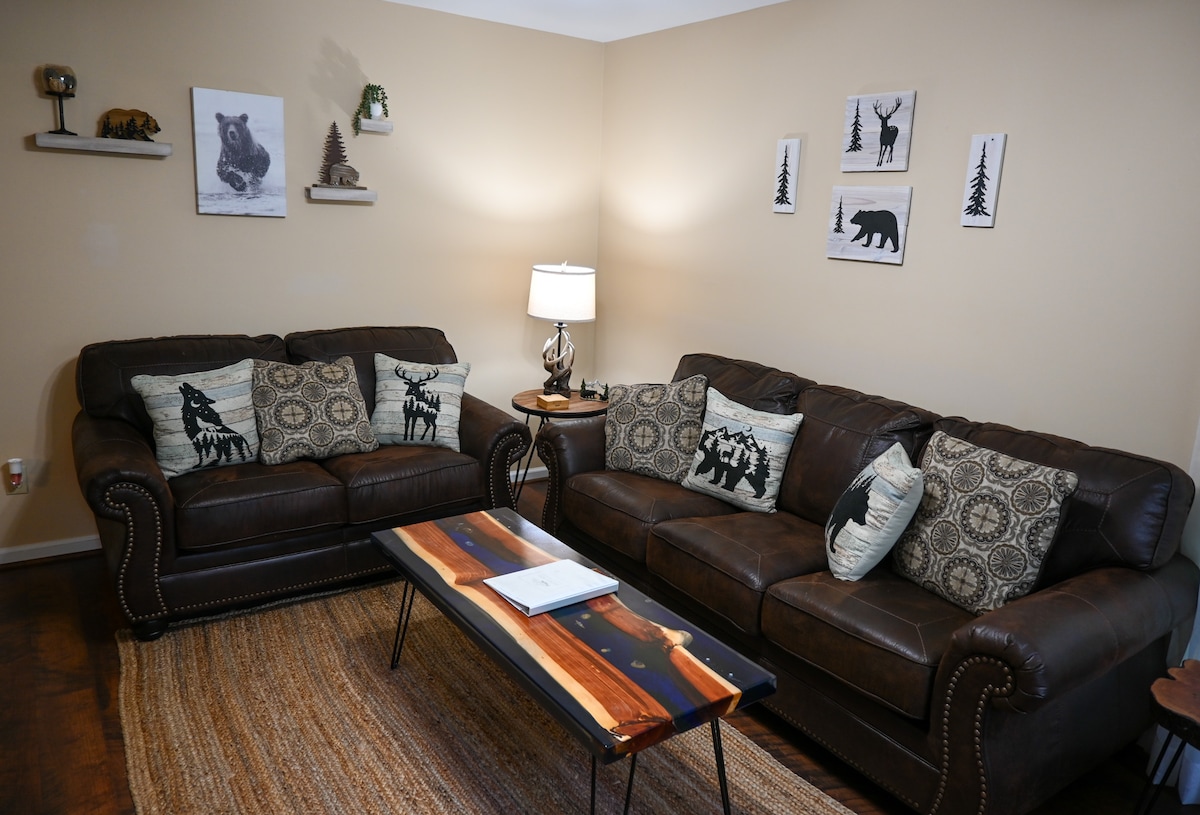 A cozy living area features two dark leather sofas adorned with decorative pillows. A rustic coffee table sits centered on a woven rug, and a lamp provides soft lighting. Artwork of wildlife decorates the walls, enhancing the cabin-like ambiance.