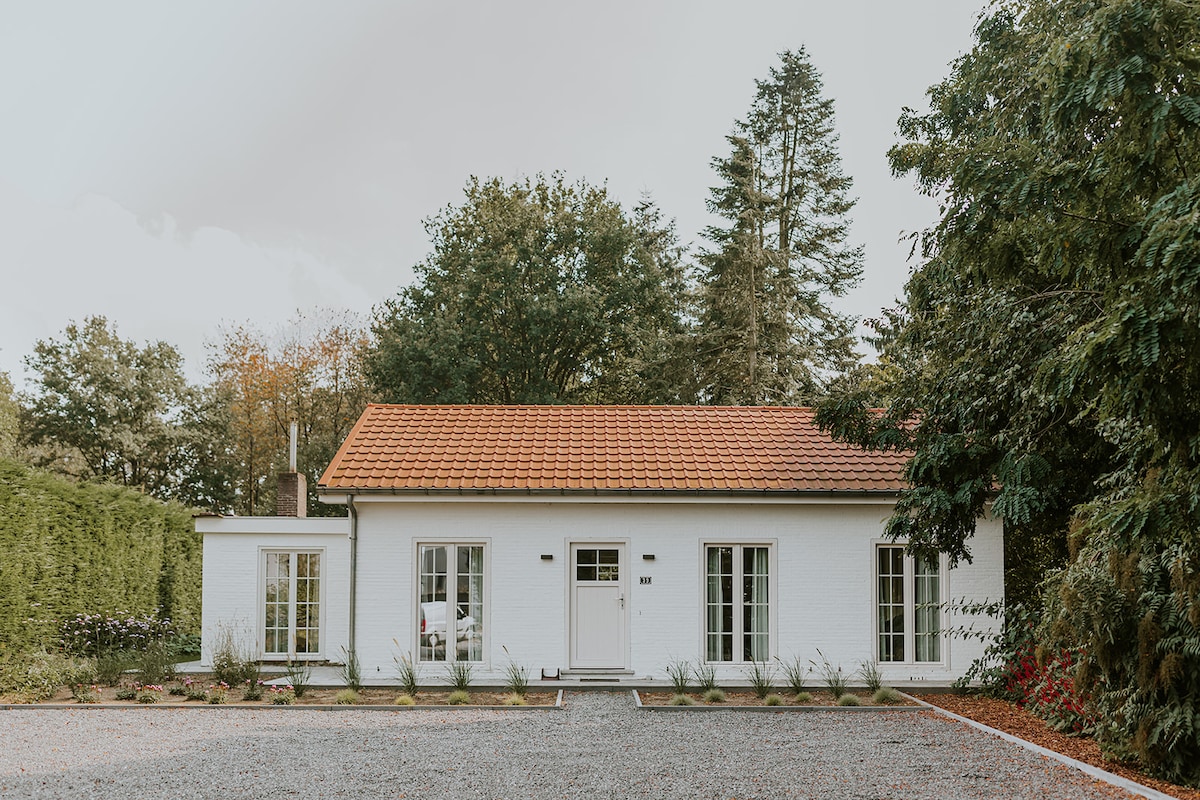 A charming single-story house features a white exterior and a red-tiled roof. Large windows on either side of the front door allow natural light to enter. The surrounding gravel driveway is lined with low-maintenance plants and bordered by greenery, creating a peaceful ambiance.
