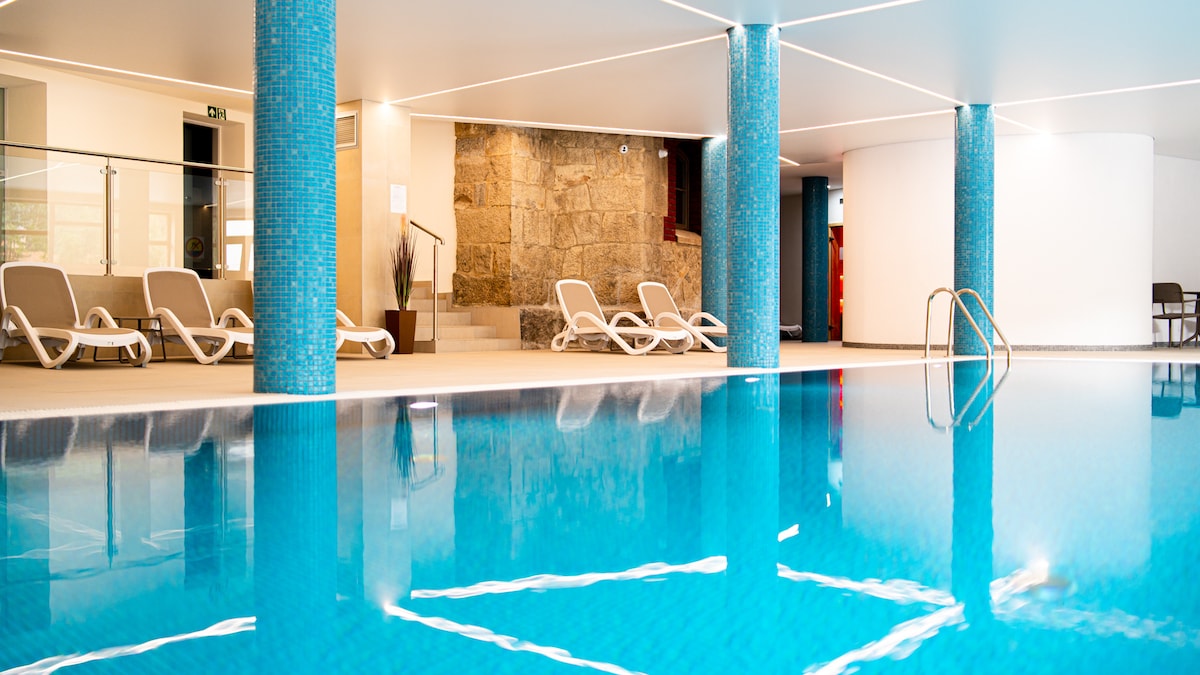 An indoor swimming pool area is featured, with clear blue water reflecting the serene environment. Lounge chairs are positioned beside the pool, and a stone wall adds texture to the backdrop. Soft overhead lighting illuminates the space, creating a calming atmosphere for relaxation.