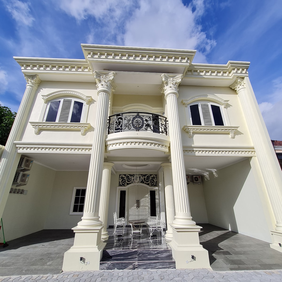 The exterior of the villa is showcased, featuring grand columns and intricate balconies. A covered patio area contains a small seating arrangement, surrounded by clean architectural lines and smooth stone flooring. The sky above is clear with soft clouds, enhancing the villa's elegant appearance.