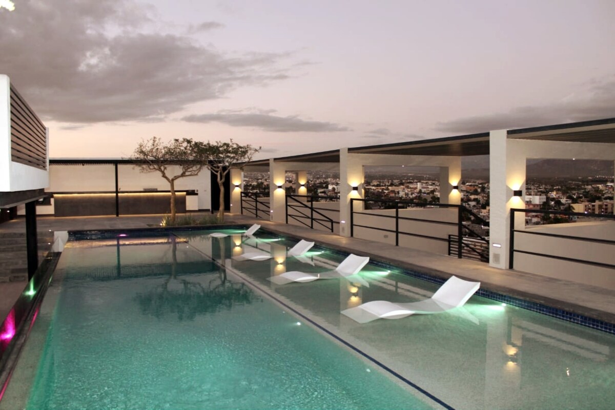 A rooftop pool area is presented, featuring a tranquil pool with illuminated edges. Several white lounge chairs are positioned around the pool, complemented by soft lighting that enhances the view of the surrounding area and the horizon.