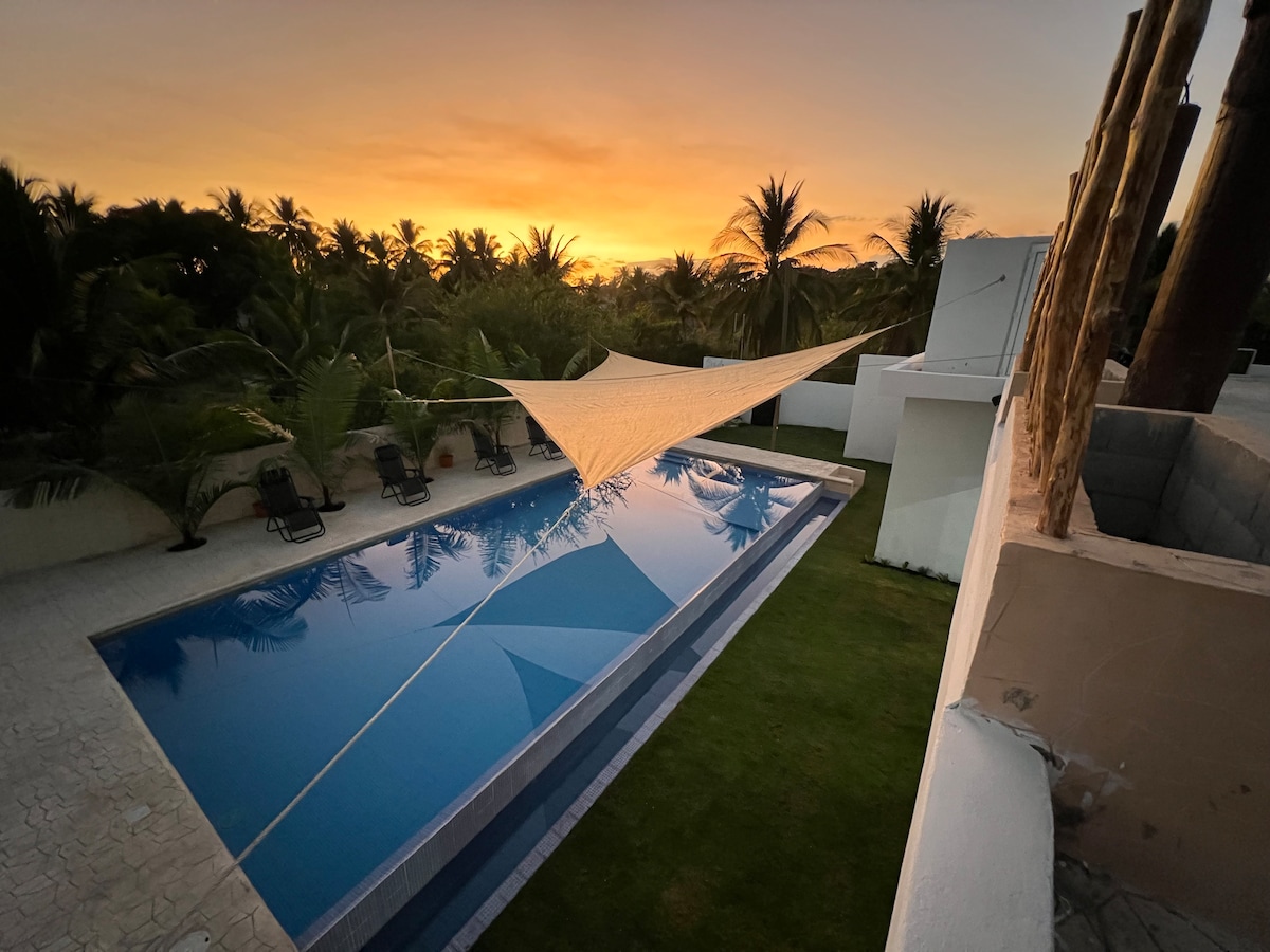 A tranquil pool area is depicted at dusk, featuring a freshwater pool reflecting the warm hues of the sunset. Lounge chairs line the poolside, accompanied by a large white sunshade providing comfort. Lush greenery surrounds the space, enhancing the serene atmosphere.