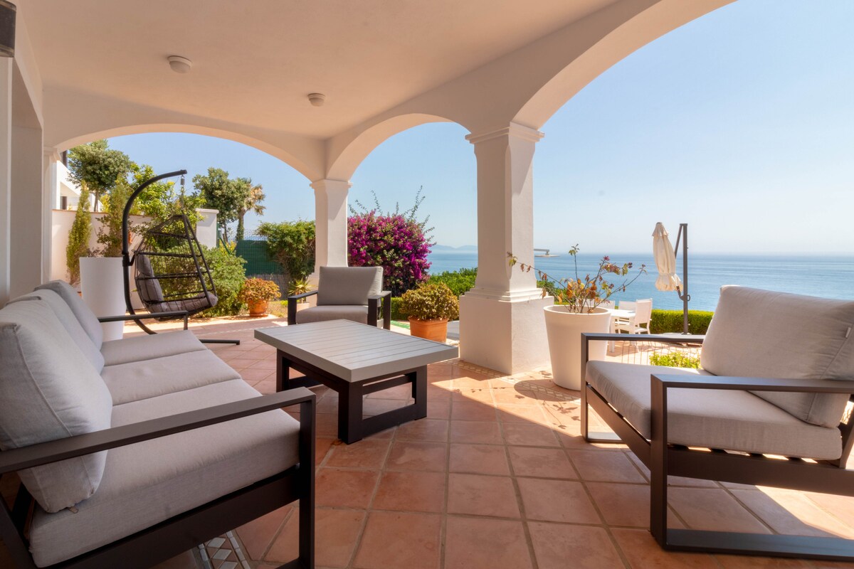 A covered terrace offers a comfortable seating area with light grey armchairs and a sleek coffee table. Potted plants and vibrant flowers add color, while a swinging chair enhances relaxation. The view stretches out toward the sea, creating a serene coastal ambience.