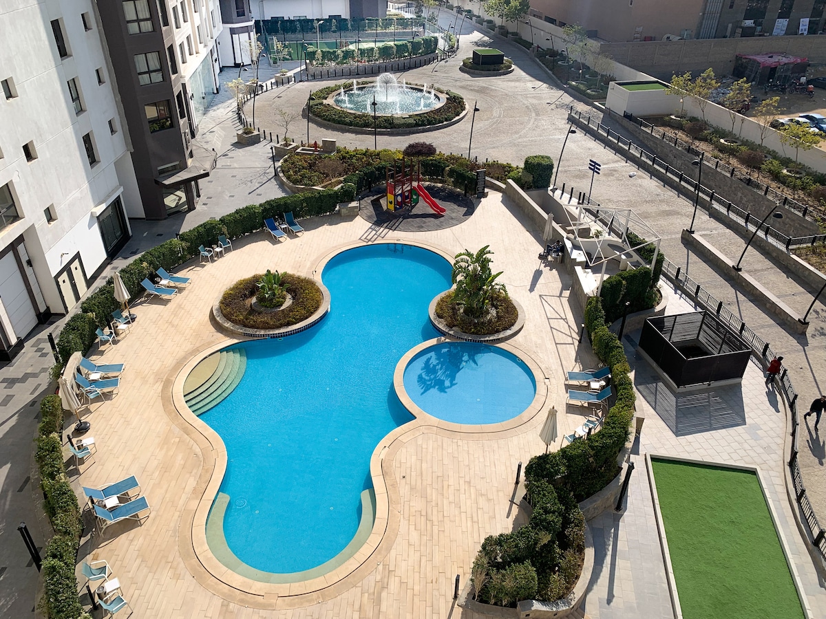An outdoor pool area is depicted, featuring a uniquely shaped pool surrounded by lounge chairs and manicured greenery. Nearby, a children's play area with slides and a fountain can be seen, with well-maintained walkways connecting the spaces.