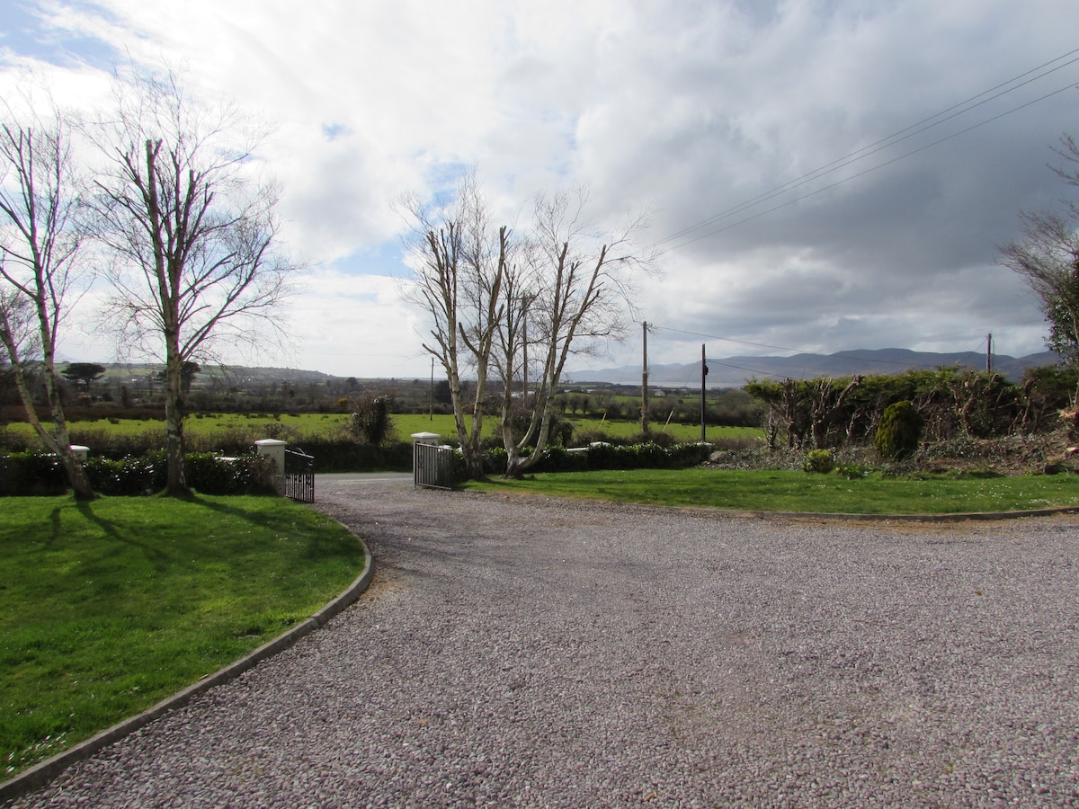 The view features an open landscape with rolling fields extending into the distance. A gravel driveway is lined with trimmed bushes, and a pair of trees stand in the foreground, complementing the serene countryside atmosphere under a partly cloudy sky.