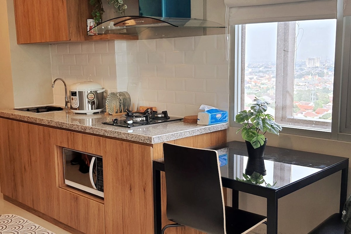 A modern kitchen area features wooden cabinetry and a granite countertop. A gas stove and kettle are visible, alongside a small table with a black chair and a potted plant. A window allows natural light to fill the space, showcasing a city view.