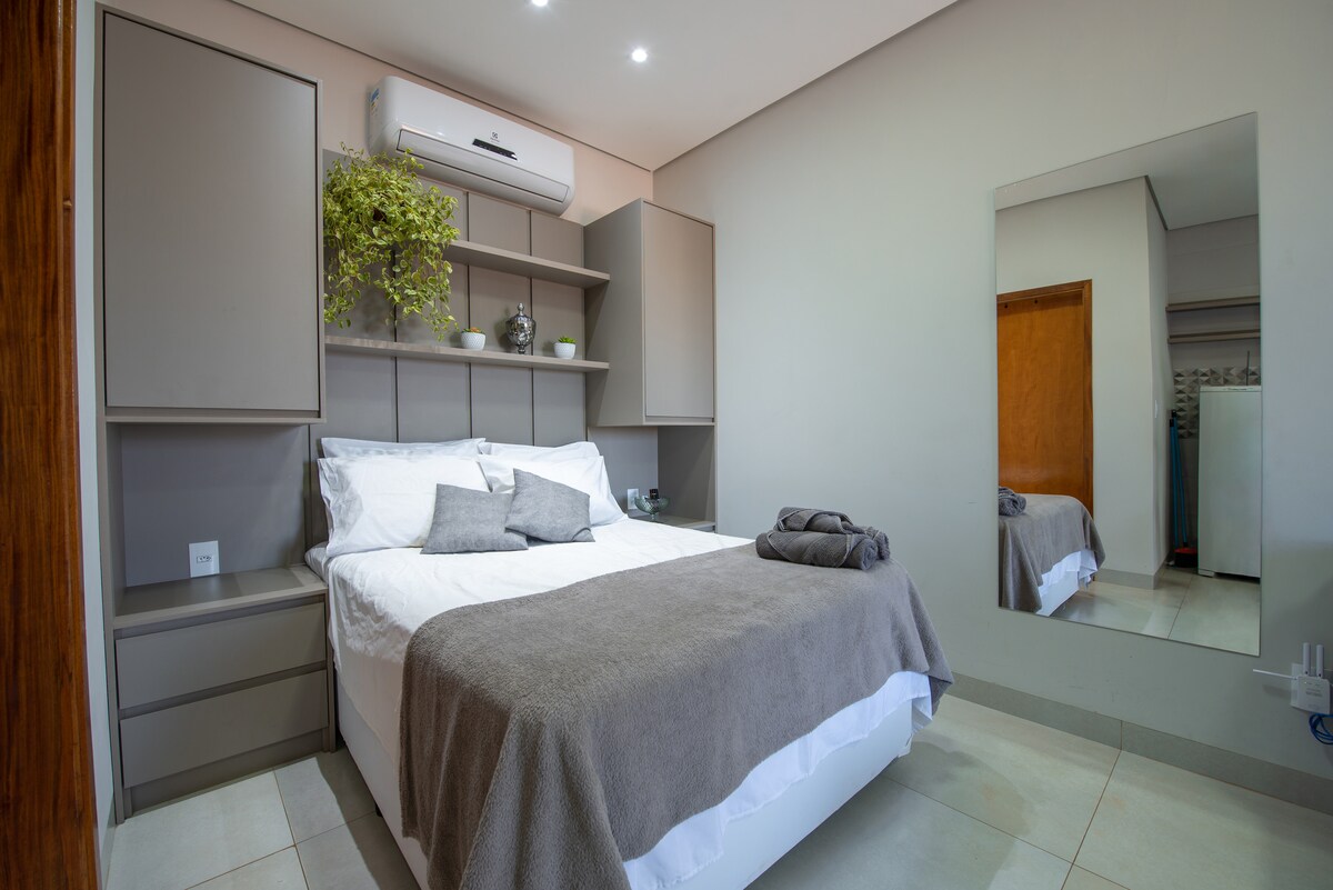 The sleeping area features a spacious bed dressed in neutral linens, flanked by bedside tables. A large mirror reflects light into the room, and a plant is placed on a shelf above the bed. Air conditioning is installed on the wall for comfort.