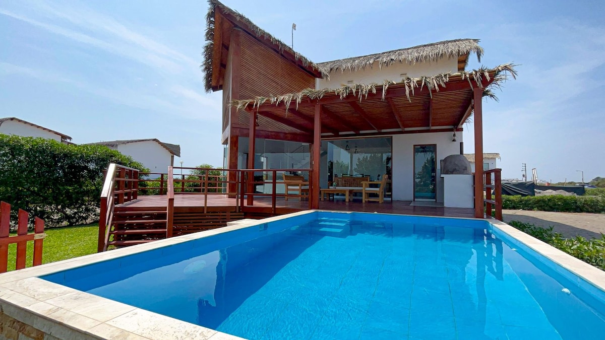 A private pool reflects the clear blue sky, surrounded by well-maintained grass and a wooden deck. The structure features a thatched roof and large glass doors, creating a seamless connection between indoor and outdoor spaces.