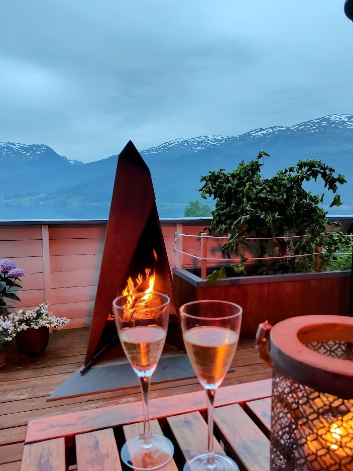 A charming outdoor terrace is highlighted by a modern fireplace, featuring a cozy flame surrounded by potted plants. Two glasses of sparkling beverage rest on a wooden table, with a scenic view of the mountains in the background, partially shrouded in cloud.
