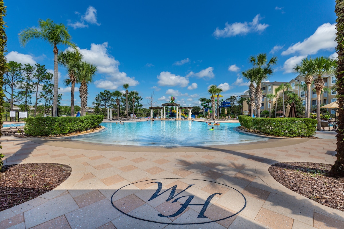 A large pool is surrounded by lush palm trees and manicured landscaping, inviting relaxation. Water slides and play areas are visible in the background. The sky is clear with scattered clouds, enhancing the bright atmosphere of the resort.