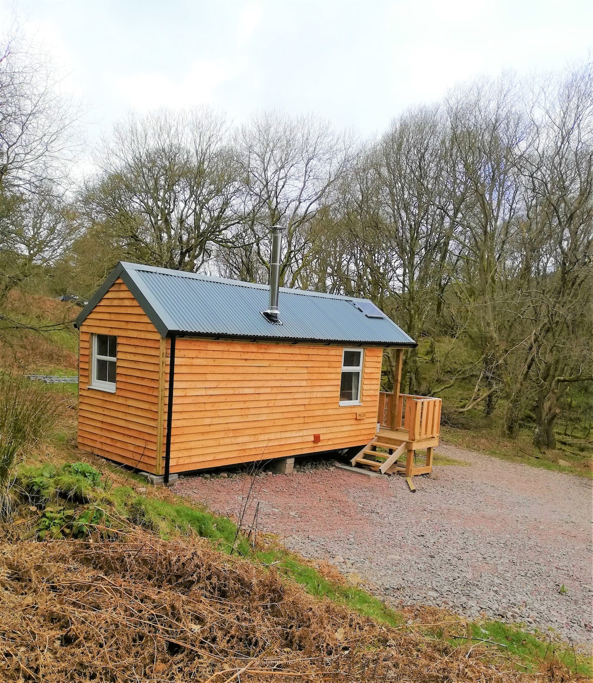A rustic wooden cabin is positioned among trees, featuring a sloped metal roof and a staircase leading to a raised deck. A wood burner pipe is visible on the roof, and gravel offers parking space in front. Natural surroundings create a serene atmosphere.
