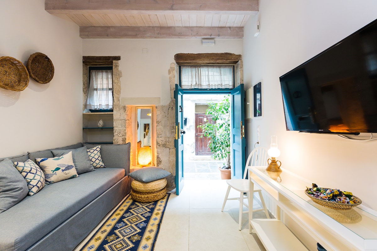 A spacious sitting area features a comfortable gray sofa adorned with patterned cushions. Light enters through open doors leading to a courtyard, enhancing the bright, airy atmosphere. A television is mounted on the wall opposite a decorative table, while natural textures are added with woven baskets.