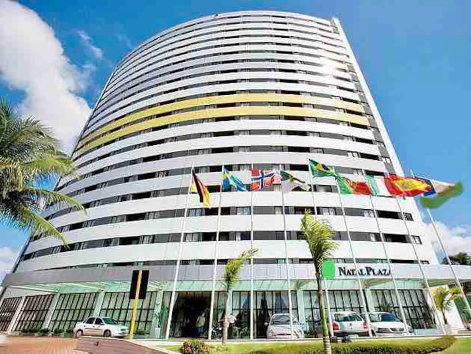The modern multi-story building features a curved design with large glass windows that reflect the sky. A row of international flags is displayed prominently in front, while palm trees line the entrance, contributing to the vibrant outdoor setting.