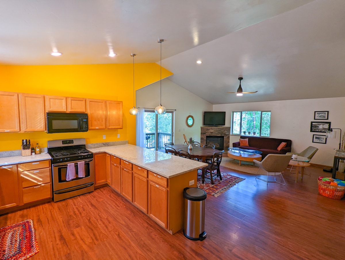 A spacious kitchen features wooden cabinetry and granite countertops, with modern appliances visible. A dining area is situated next to a large window, allowing natural light. The cozy living space includes comfortable seating and a view of the outdoors, enhancing the welcoming atmosphere.