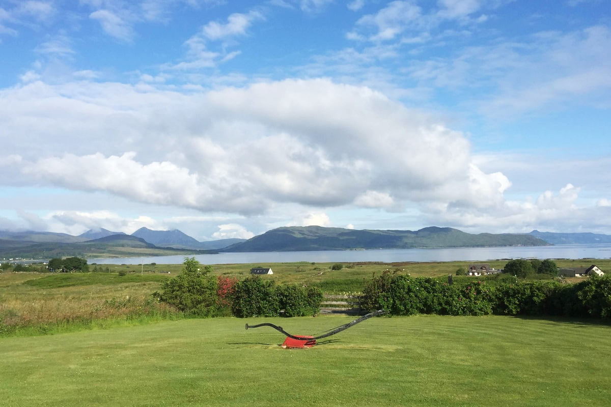 A broad expanse of green lawn extends across the foreground, leading to a panoramic view of the Red Cuillin mountains and Broadford Bay. Fluffy white clouds drift in a blue sky, reflecting the tranquil landscape that surrounds the location.