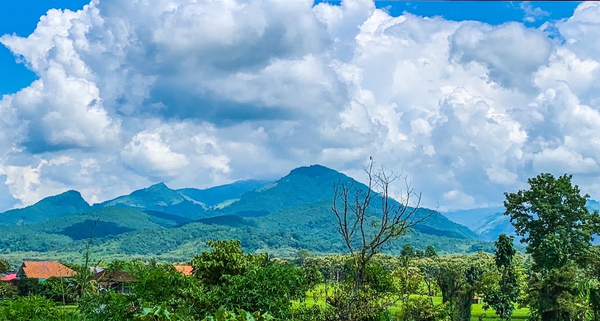 A scenic view captures rolling green mountains under a vibrant sky filled with clouds. Lush greenery is visible in the foreground, complemented by a few trees and distant rooftop structures, creating a peaceful and natural landscape.