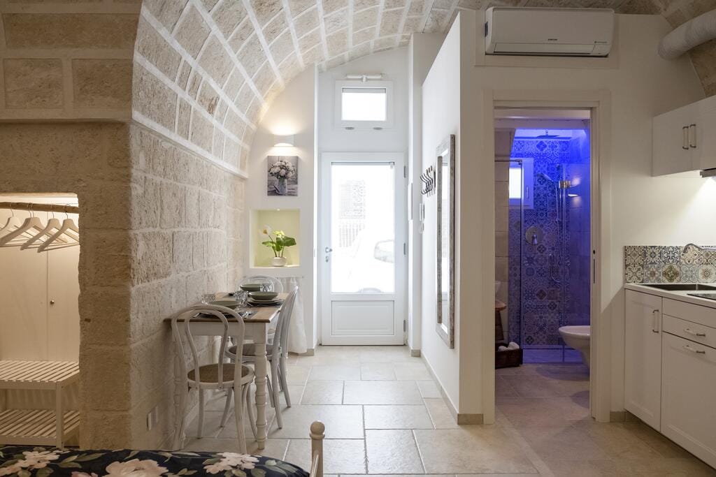 The interior of the studio features high arches and cream-colored stone walls. A small dining area is positioned near the entrance, while a modern kitchen is visible, complete with cabinets and appliances. A bathroom with a colorful tiled shower is adjacent, enhancing the space's functionality.