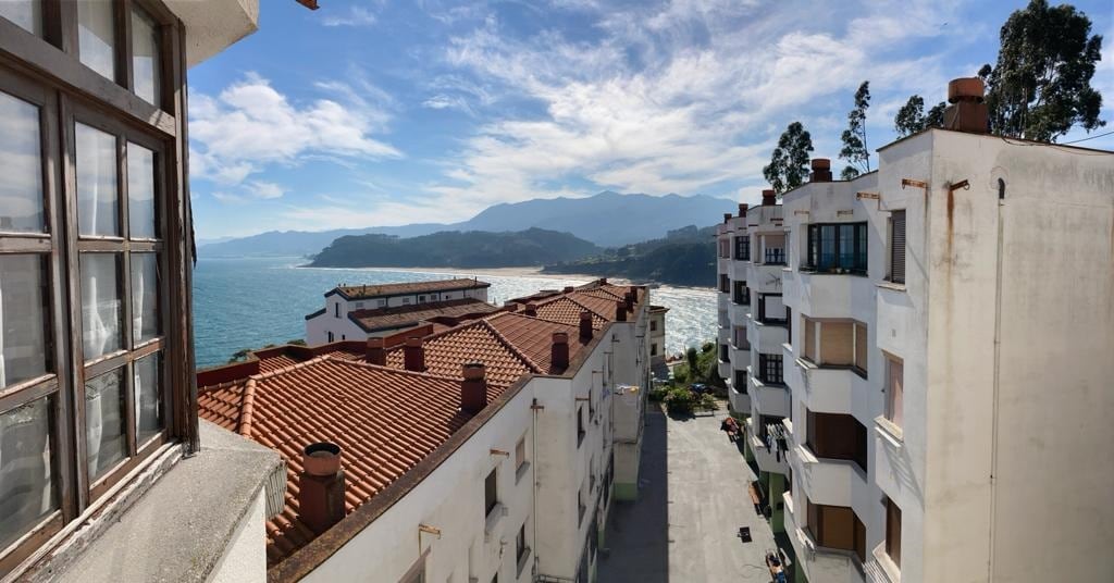 A scenic view reveals rooftops of neighboring buildings leading towards the coastline. The ocean glistens under sunlight, with distant mountains framing the horizon. Soft, fluffy clouds drift in a bright blue sky above, enhancing the coastal ambiance.