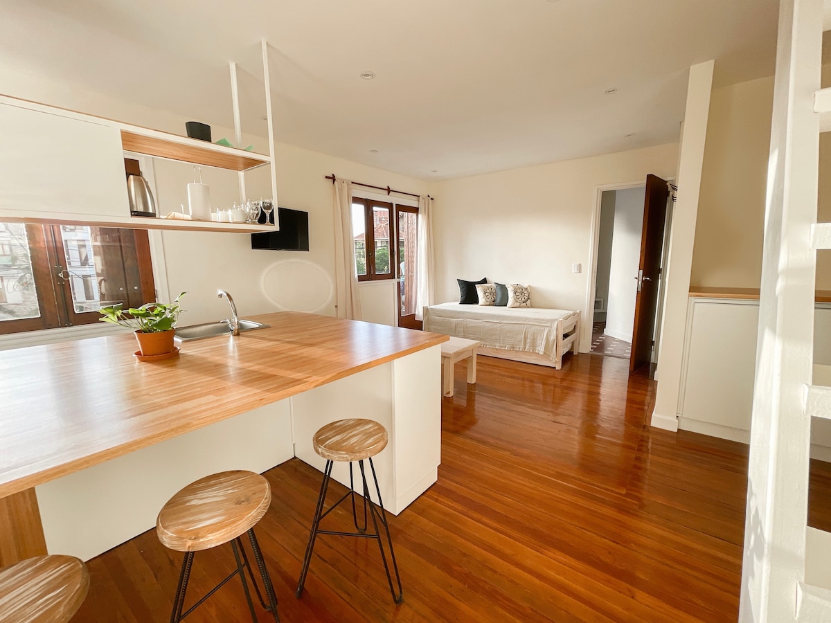 A modern kitchen area features a wooden countertop with three round stools and a sink. Natural light fills the space from large windows. A sofa bed is situated in the living area, complemented by decorative pillows. Wooden flooring creates a warm atmosphere.