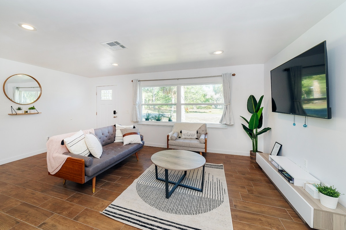 A spacious living area features a stylish gray sofa adorned with decorative pillows, complemented by a cozy chair. Natural light fills the room through large windows, highlighting the wood-inspired floor and modern decor. A round coffee table and a flat-screen TV complete the inviting ambiance.