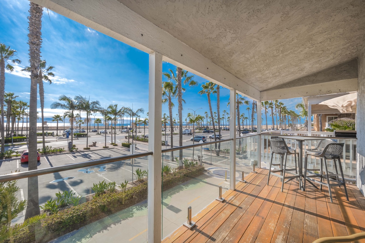 A spacious balcony features wooden flooring and clear glass railings, providing unobstructed views of palm trees lining the beach and the ocean beyond. A modern outdoor dining set is visible, creating an inviting space to enjoy the coastal scenery.