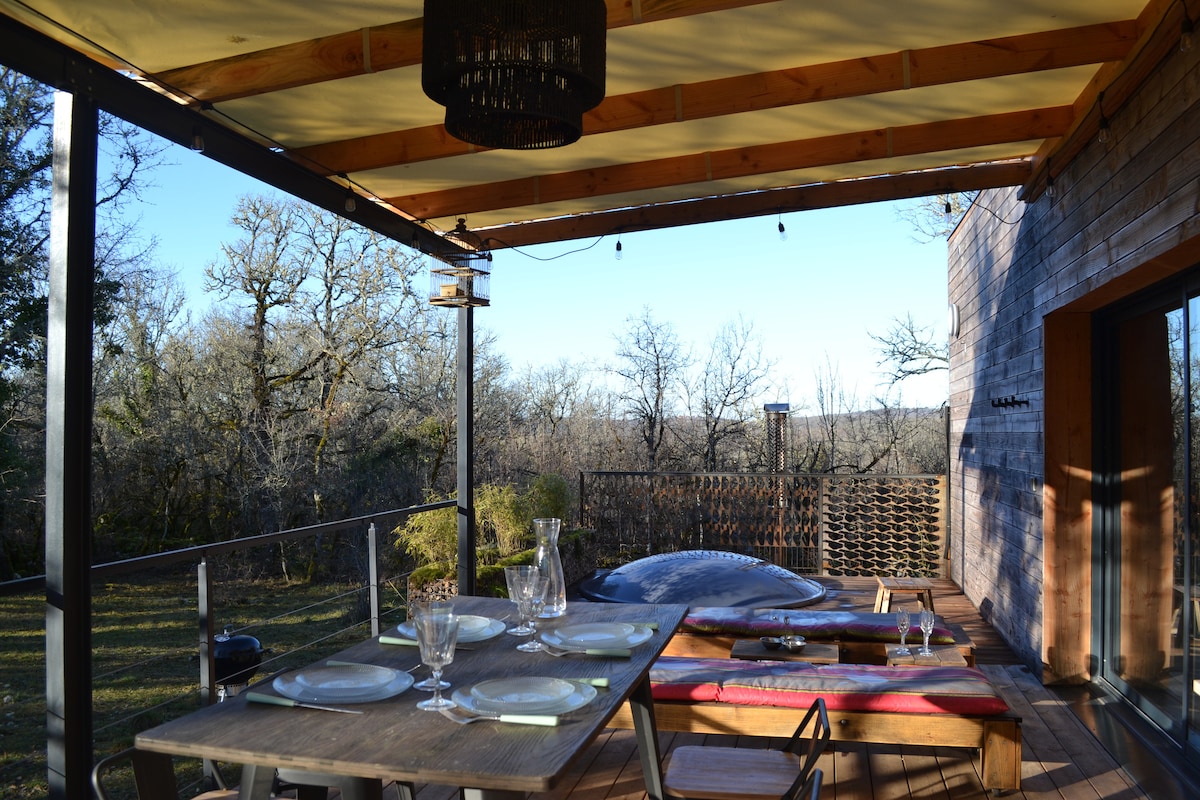 A covered terrace features a wooden dining table set with glassware, overlooking a serene natural landscape. A cozy seating area with a colorful cushion invites relaxation, while a Nordic bath is positioned nearby, providing an opportunity to enjoy the outdoors.