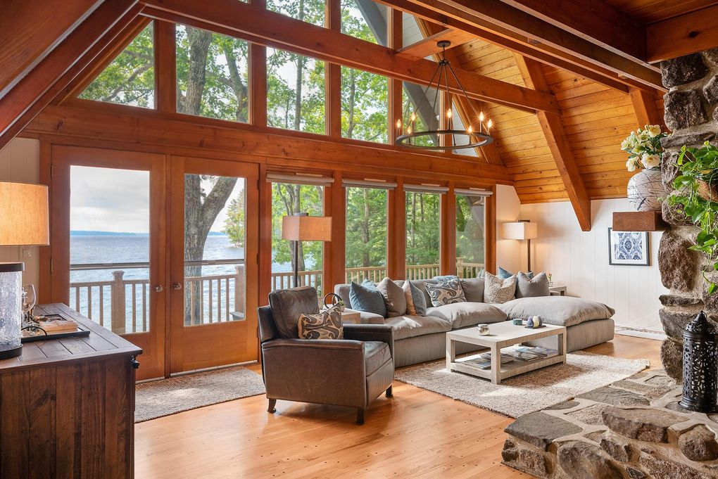 A welcoming living area features vaulted wooden ceilings and large glass doors that open to a lakeside view. A plush sectional sofa, adorned with cushions, is positioned around a low coffee table on a soft area rug. A stone fireplace adds a rustic charm to the space.