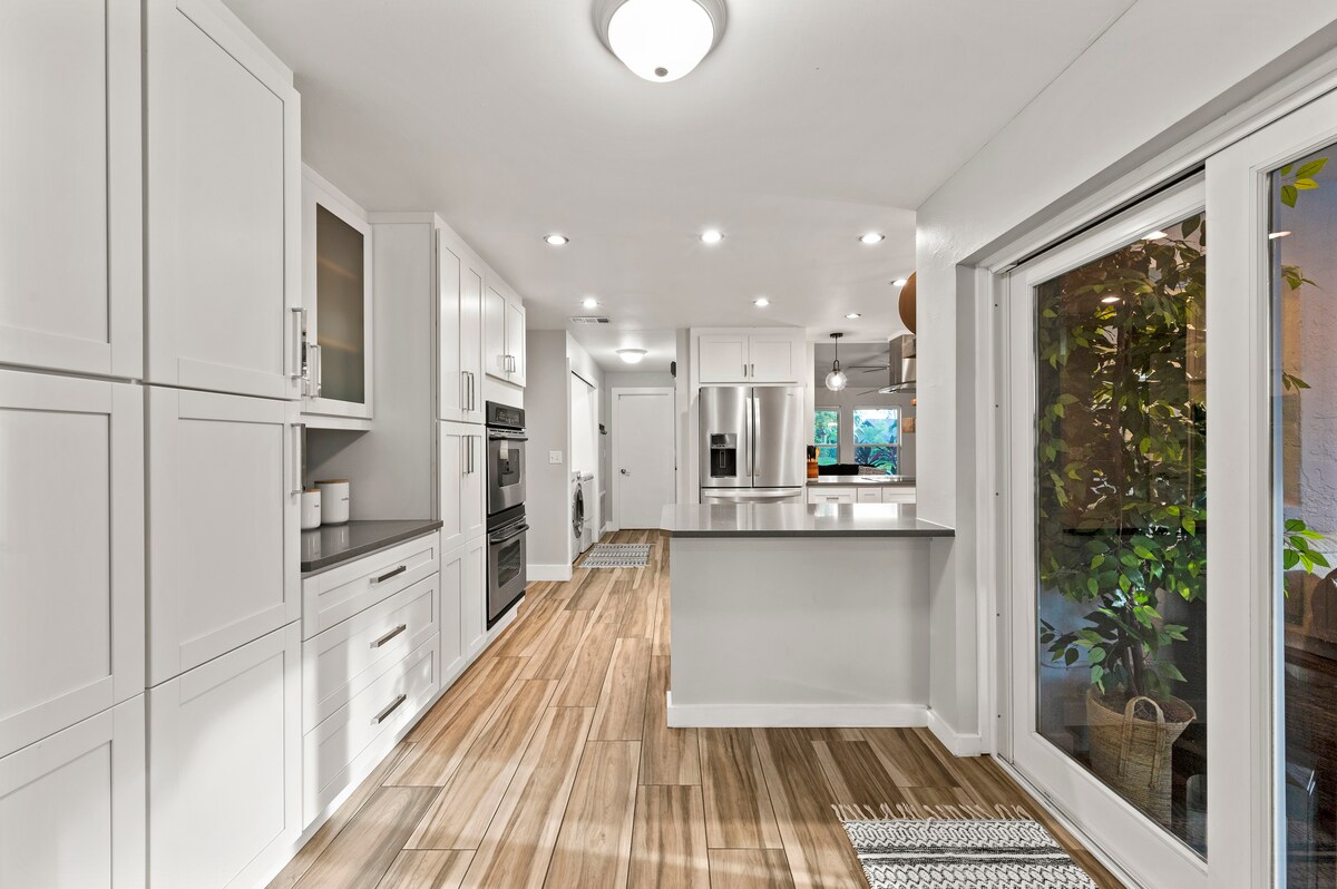 A modern kitchen is presented with bright white cabinetry and sleek appliances. Ample counter space is available for meal preparation. Natural light streams in through a large glass door, offering a view of the outdoor space. Warm wood flooring adds a cozy touch.