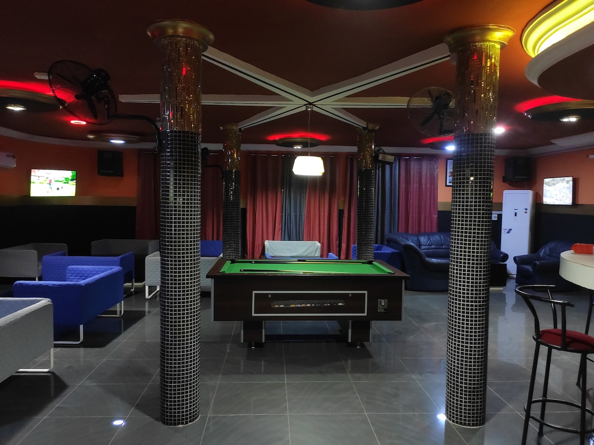 An entertainment area is shown, featuring a billiards table at its center. Surrounding seating options include comfortable couches and chairs in various styles. The walls are decorated with dark curtains, and ceiling fans can be seen for airflow. Soft lighting enhances the welcoming environment.