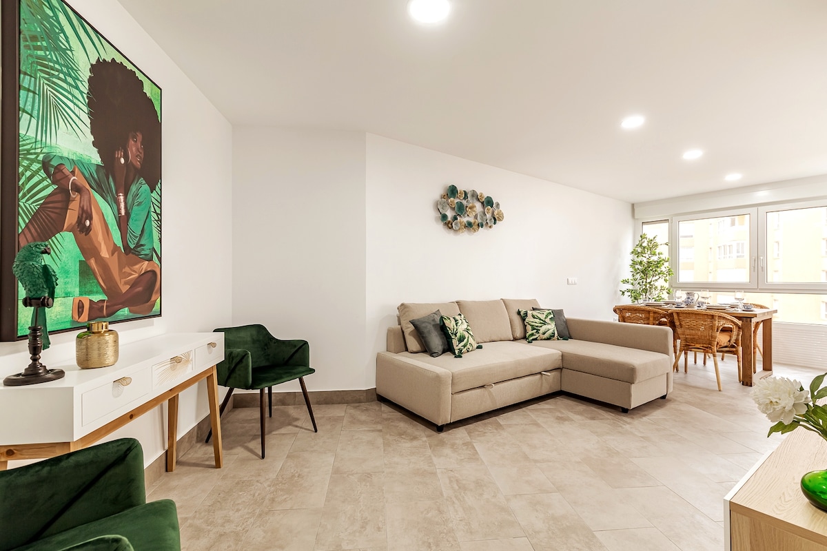 A spacious living area features a comfortable sectional sofa in neutral tones, paired with a green armchair. A dining table with wooden chairs is positioned near panoramic windows, allowing natural light to fill the room. A vibrant wall art adds a touch of color to the decor.