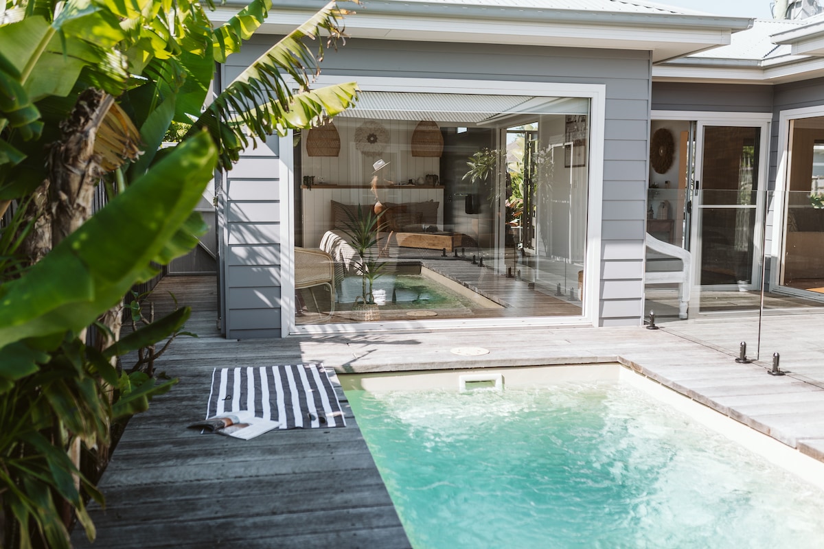 A private plunge pool is observed beside a wooden deck, surrounded by lush tropical plants. Glass doors open to an inviting living space, creating a seamless transition between indoor and outdoor areas. A striped towel lies on the deck, accenting the relaxed atmosphere.